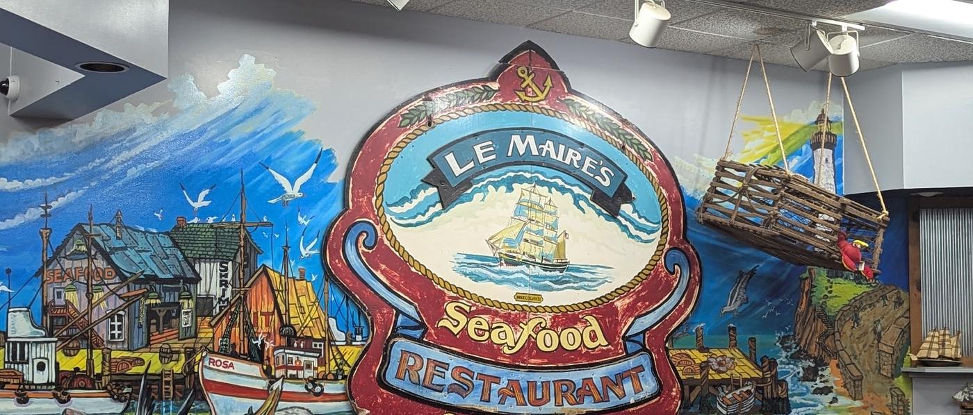 The carved LeMaire's Seafood Restaurant and Market sign, with a tall ship and an anchor, mounted on a hand painted harbor mural of fishing shacks, boats and a lighthouse.